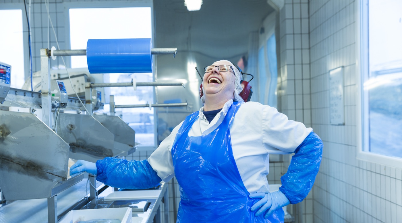 Glad medarbeider i blått forkle i et fiskeforedlingsanlegg